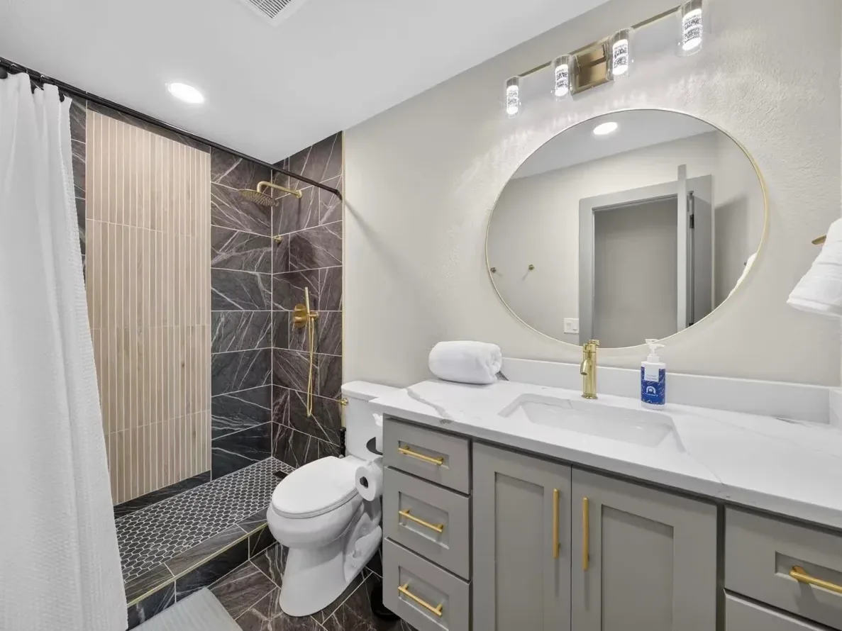 Piedmont bathroom remodeled by MASB: walk-in shower in dark charcoal marble-look tile with a wood-tone slat accent wall, a gray shaker vanity with gold pulls, and a round gold-framed mirror