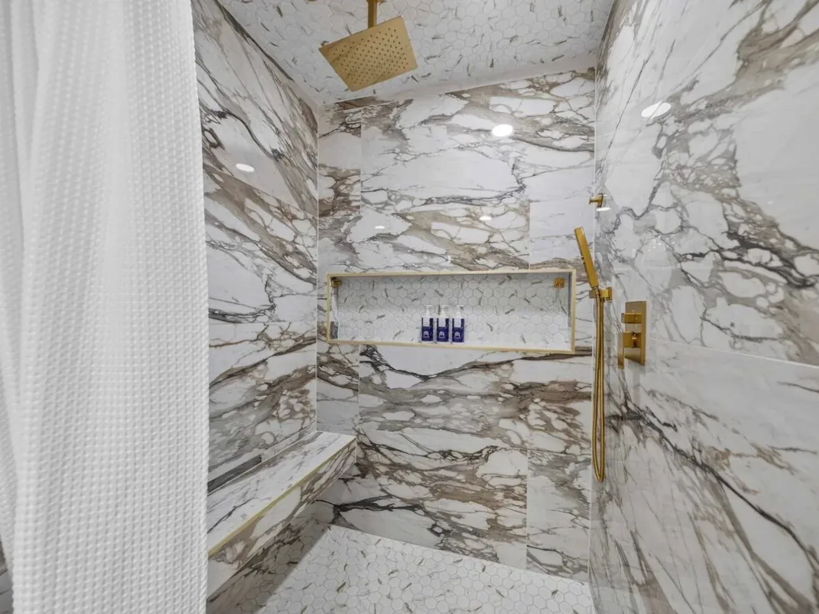 Walk-in shower in a Piedmont bathroom remodel with white-and-gold marble-look tile, a built-in bench, a long gold-trimmed niche, and a square gold rain head