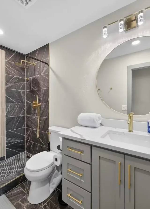 First bathroom: walk-in shower in dark marble with a wood-slat accent wall, gray vanity with gold pulls, and a round gold mirror