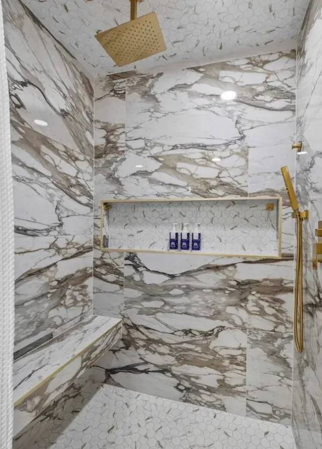 Walk-in shower with a built-in bench, gold-trimmed niche, square gold rain head, and handheld shower in white marble